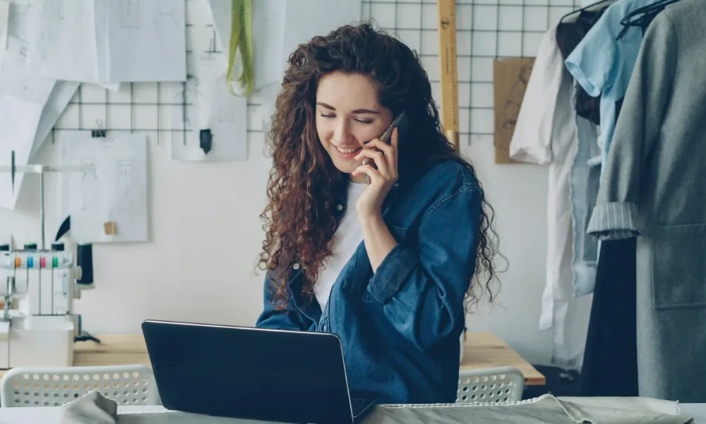 A shop owner on the phone at her laptop, smiling, between the clothes rails of her workshop